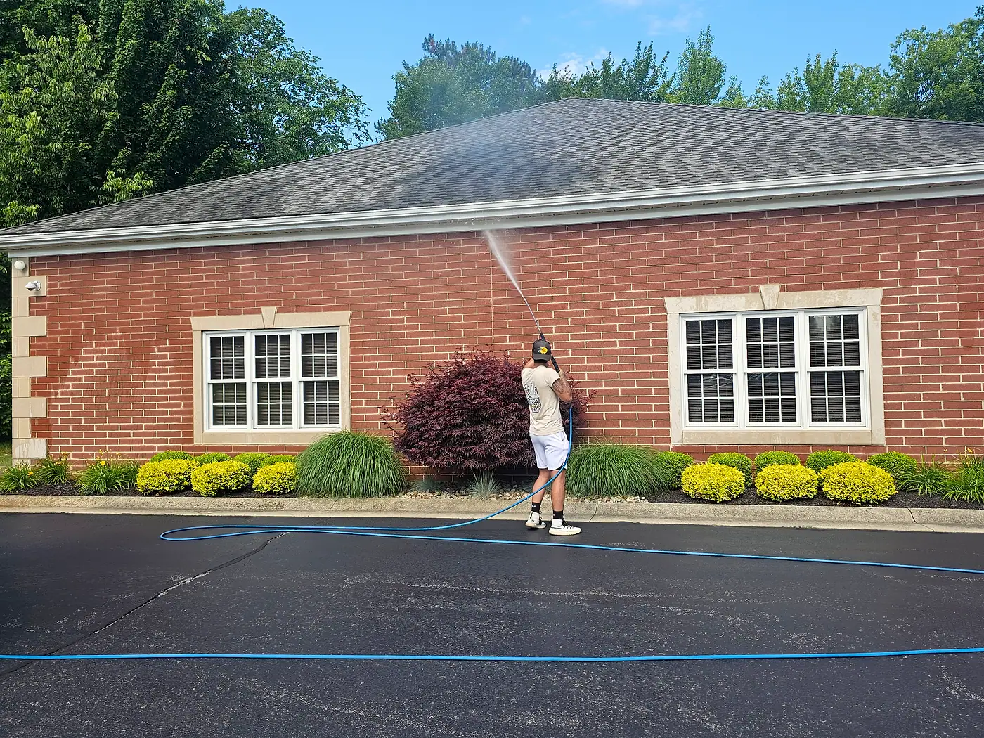 Worker pressure washing the lot beside a brick office building with flowering shrubs