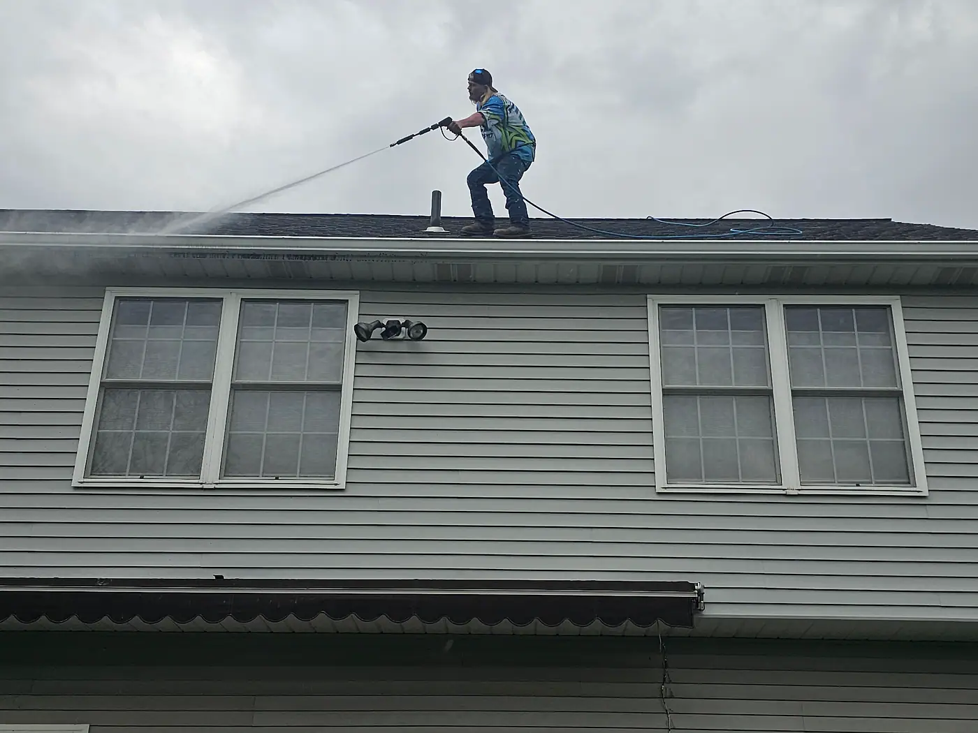 Worker at the roof edge of a gray two-story house cleaning the gutters