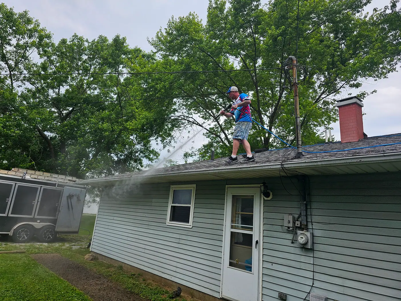 Worker on a low roof cleaning gutters with trees overhead