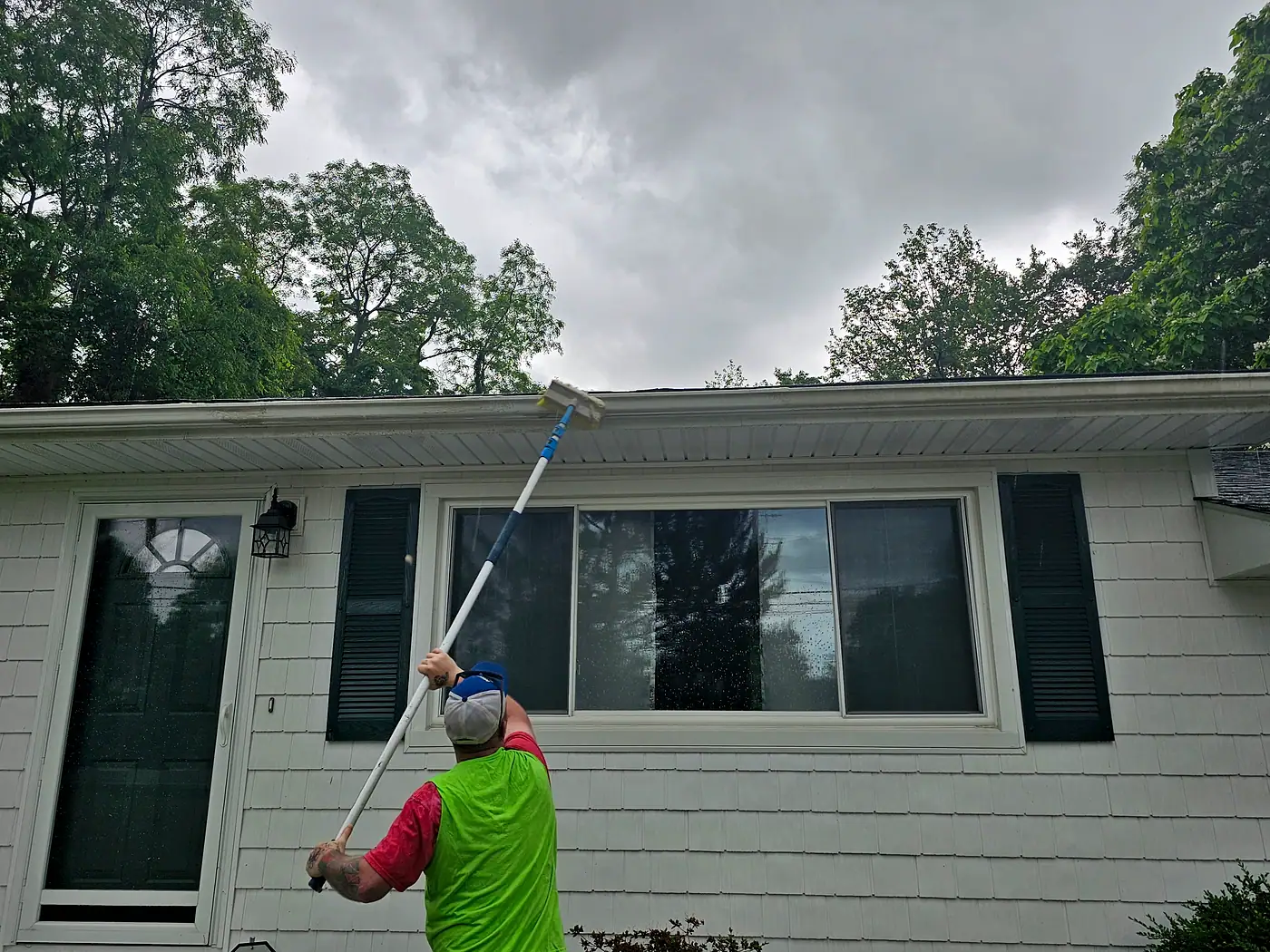 Worker using a pole to clean the gutters along the front of a gray house