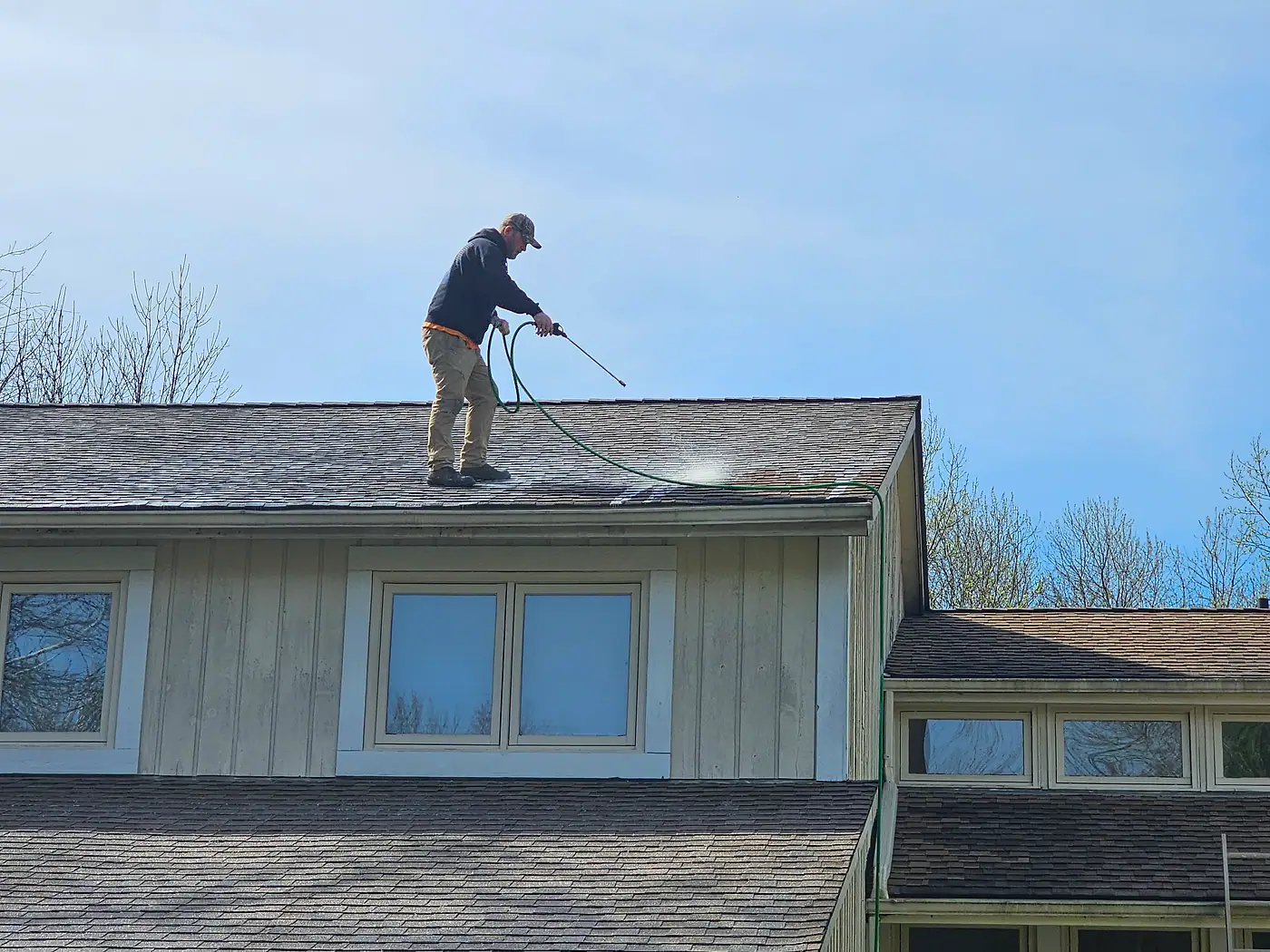 Worker standing on a shingle roof cleaning it under a clear blue sky