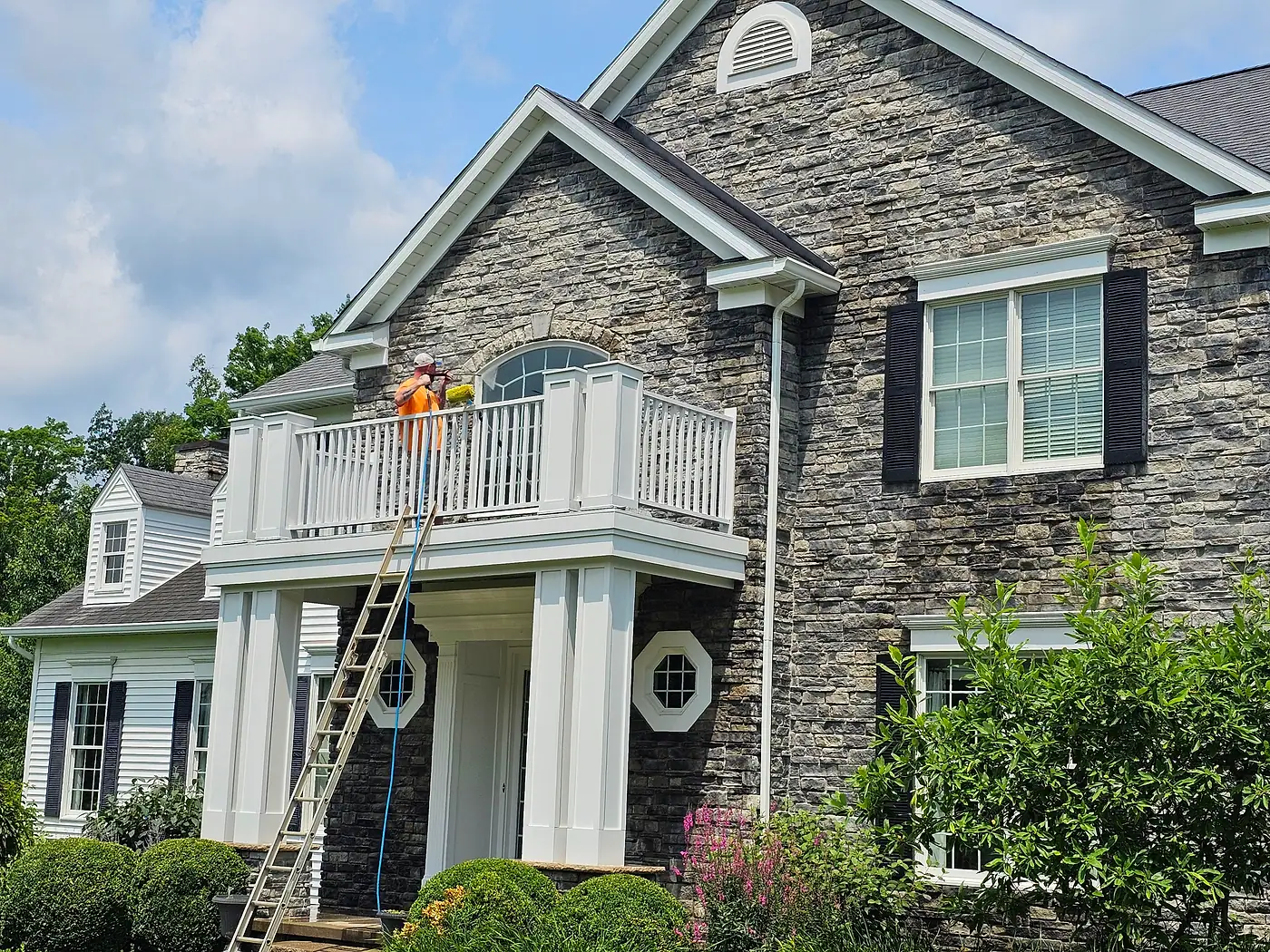 Crew member washing a stone-front home from a balcony, with a ladder and hose, in Chagrin Falls