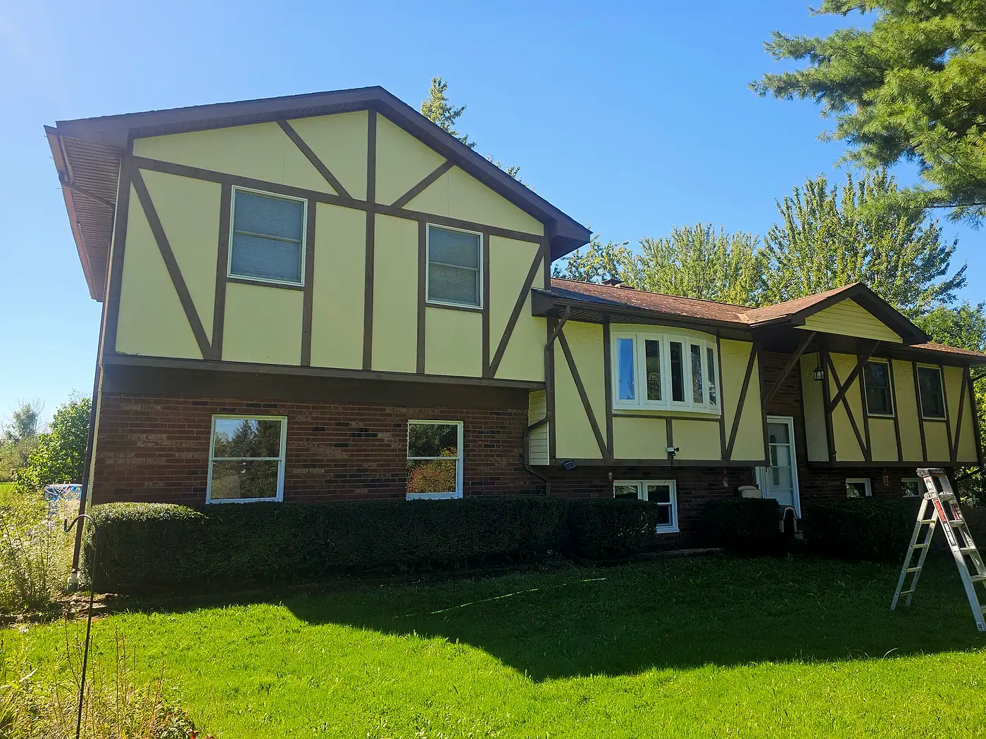 Home with a brick base, pale yellow panels, brown trim, and a ladder against the eaves in Chardon