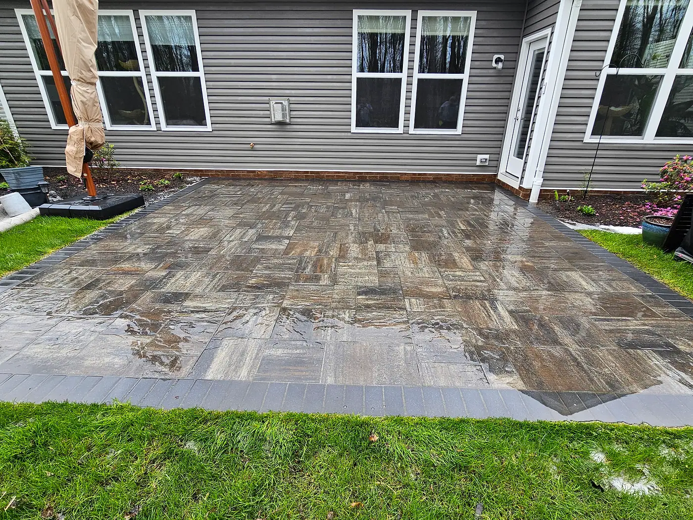 Brown and gray paver patio with a dark border, wet from cleaning, behind a gray house in Concord