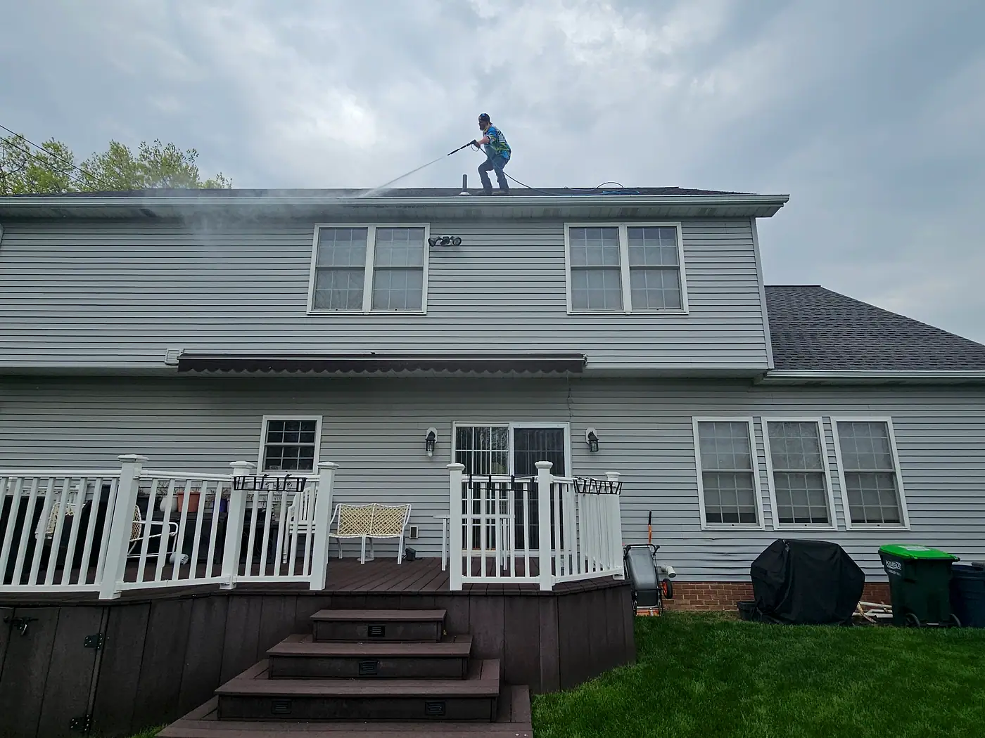 Crew member on the roof edge of a two-story home with gray siding and a deck, spraying with a pressure washer in Wickliffe